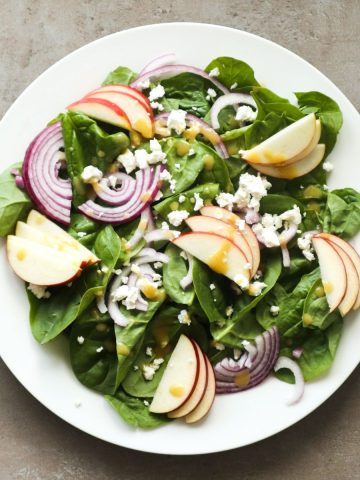 Spinach salad with feta cheese, Apple and honey mustard dressing on a white plate.