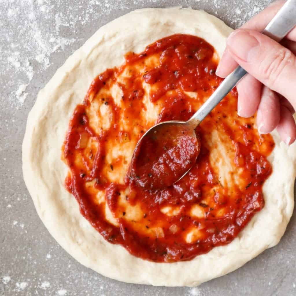 Pizza sauce spread on pizza dough with a spoon.