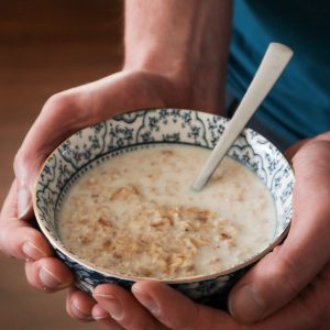 a close-up of overnight oats with yogurt and water in a delft blue bowl with two hands an a wooden background.